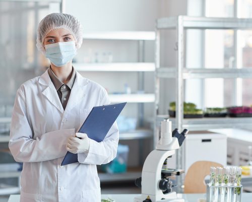 Waist up portrait of young female scientist wearing face mask and looking at camera while standing in medial laboratory and holding clipboard, copy space